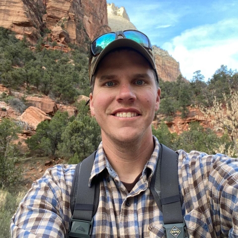 Nate grins for the camera, posing in front of a gorgeous backdrop of rocky mountains and evergreens.