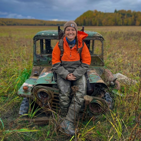 A person sits on a frame of an old jeep in the middle of a field.