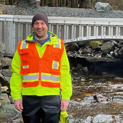 A person wearing an orange visibility vest smiles in front of a bridge with a stream running under it.