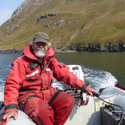 Man wearing orange float suit steers a skiff with water and island in the background.