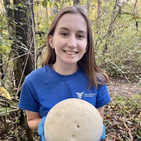 Digital Communications Intern Caitlin May stands in the woods and holds a giant puffball mushroom.