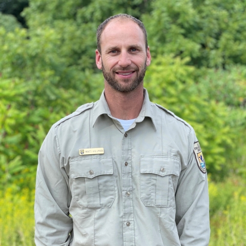 A man in a tan shirt, standing in front of some plants