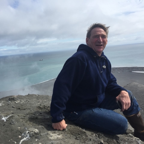 Man in sweatshirt sits on brown island with pale ocean and cloudy sky behind him.