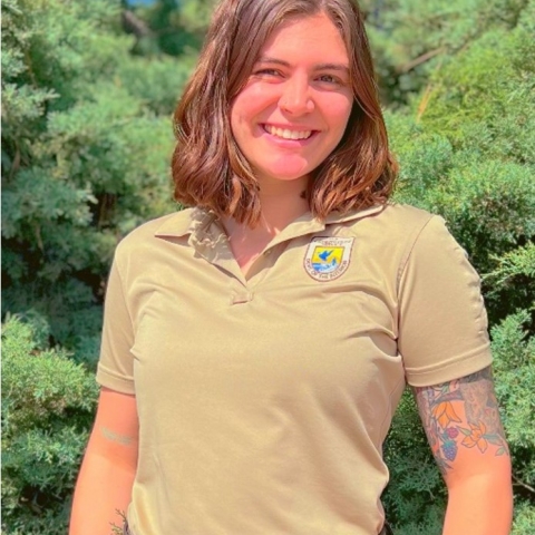 Kayla smiles in a tan FWS uniform polo in front of green pine trees