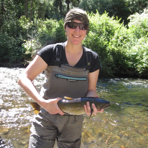 Brook Silver, Fish Biologist, wearing tan waders, black t-shirt and holding a salmonid. She is standing in a shallow creek. 