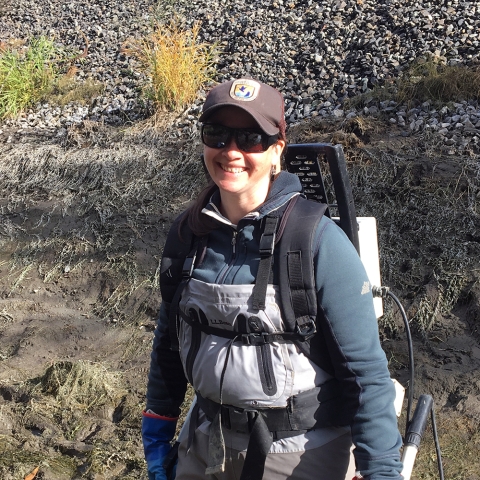 A smiling woman in sunglasses, Service ballcap, fleece jackets, and waders wears a backpackable electroshocker box on her back and holds two rods in her hands for carrying the current to the water.