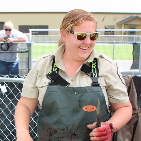 Smiling woman in sunglasses, waders, and Service uniform short-sleeved shirt holds a fish by the tail with a gloved hand.