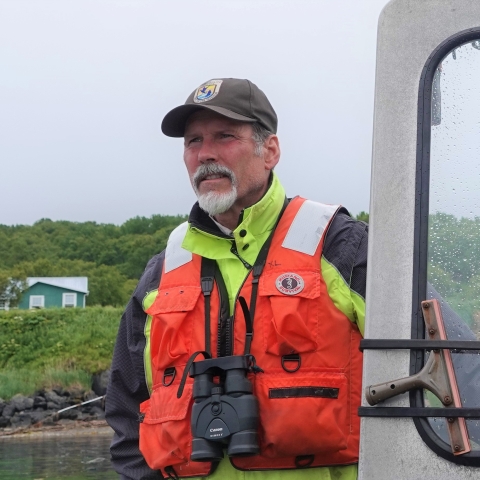 man in usfws hat and lifejacket steers a skiff with hill and houses on the shore in the background