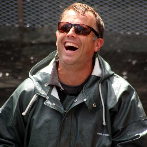 A laughing man in sunglasses and rainjacket stands up to his waist in water in a concrete tank.