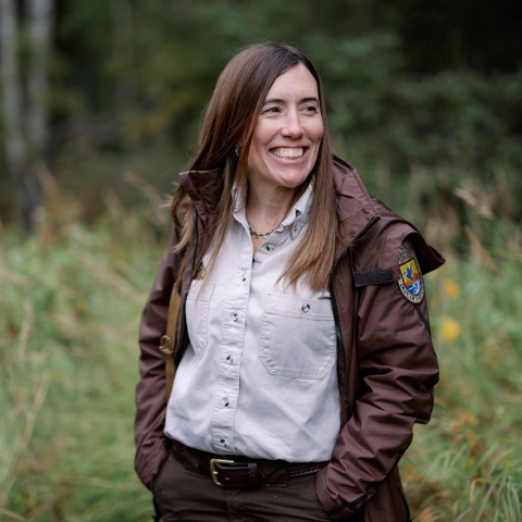 A woman with long brown hair wears a Refuges uniform brown jacket, tan shirt, and brown pants and stands outside.