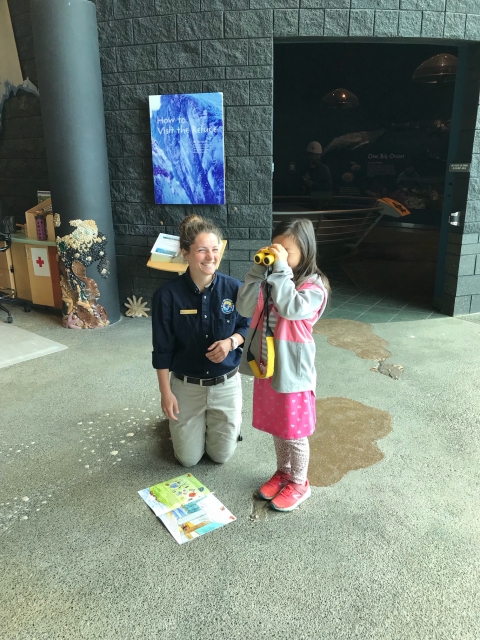 Refuge volunteer down at kid level on her knees, assisting a child with using binoculars as part of the Jr Biologist program at Alaska Maritime National Wildlife Refuge Visitor Center