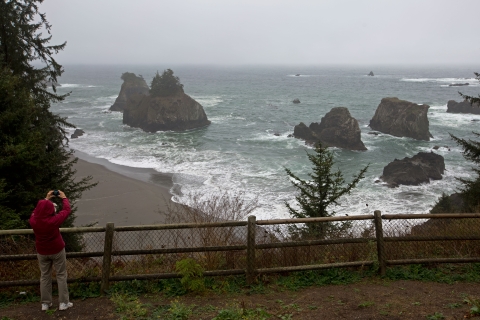 A person on a coastal cliff takes a photo of offshore rocks amid pounding surf