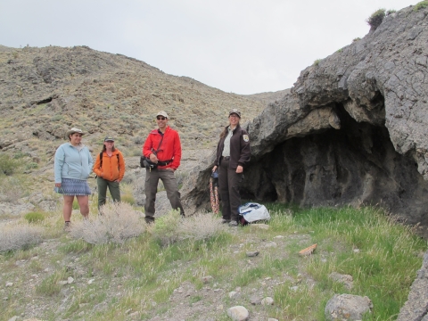 Three women and a man in hiking gear stand beside a cave and agave roasting pit in Desert National Wildlife Refuge