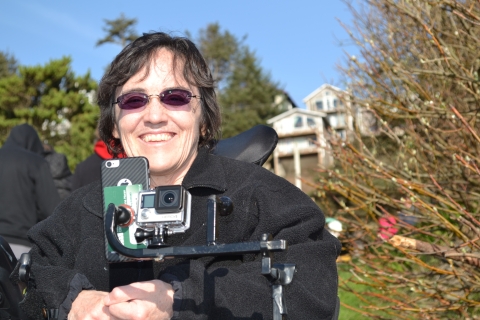 A woman in sunglasses smiles, facing camera, from her motor-powered wheelchair that she drives with her chin. Some electronic devices are visible on a bar below her face.