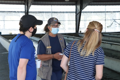 A masked, uniformed volunteer stands next to water-filled fish raceways with two visitors.