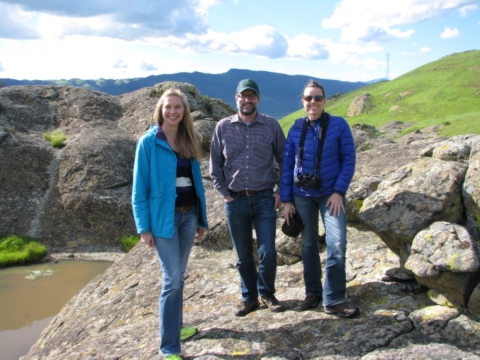 Three people standing on a large rock outcrop overlooking a pool of water with hills and mountains in the background.