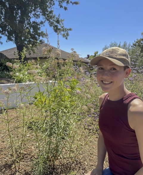 A woman wearing a maroon tank top and baseball cap sitting next to a green plant