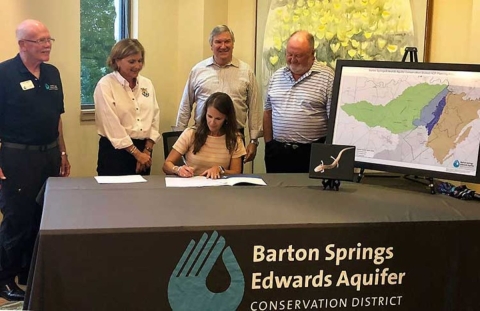 A woman sits at a table while signing a document. Four other people stand behind her and watch.