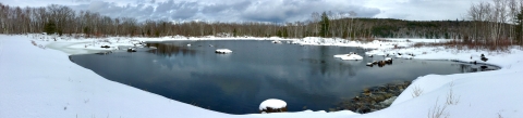 A wide-angle view of a calm dark blue pond with snow on all sides and forested hills in the distance