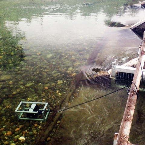 Photo of a metal frame around a large black camera set on a gravel river bottom, leashed to a bridge.