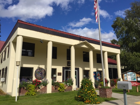 A sunny summer day with scattered small clouds. View of the front of the main hatchery building, with a row of 6 plain columns supporting a roof overhang on a 2-story structure. Blooming flowers spill out of brick planters. A flagpole with an American flag, mailbox, and informational signboard are out in front. An artwork mosaic is attached to the wall between two windows.