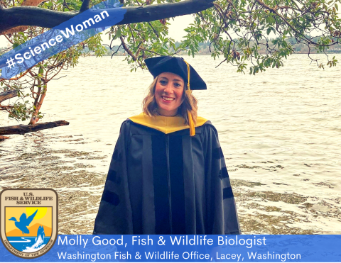 Woman in graduation attire standing in front of a body of water beneath a tree branch