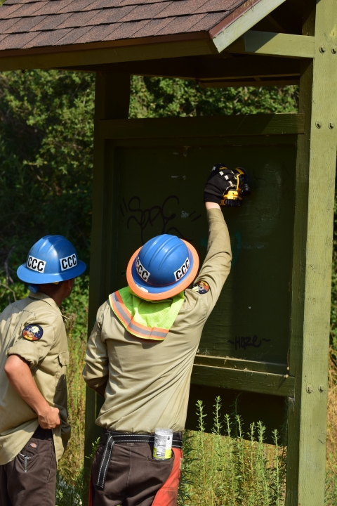 Two individuals from the California Conservation Corps standing in front of refuge kiosk to sand off graffiti.