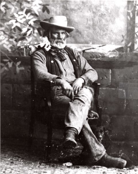 Black and white photo of a man with a beard sitting on a chair infront of a stone wall.