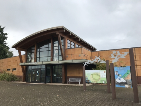 Large building with glass windows and tan siding; angled roof; sign that says "visitor center"; interpretive panels near building; stone paved ground