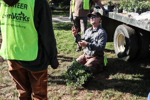 Person kneeling on the ground with several potted plants, holding one in hand, speaking to people circled around him