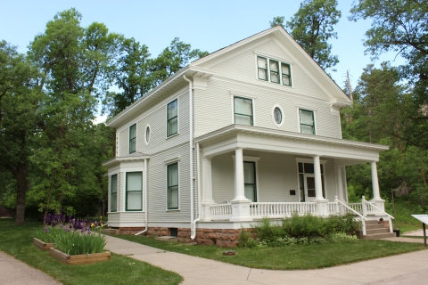 A large two story white house with green trim sits on a manicured green lawn surrounded by trees and blue sky.