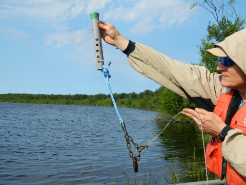 a man holds a temperature logger apparatus - a gray cylinder with a rope and chain attached - at arm's length above the water