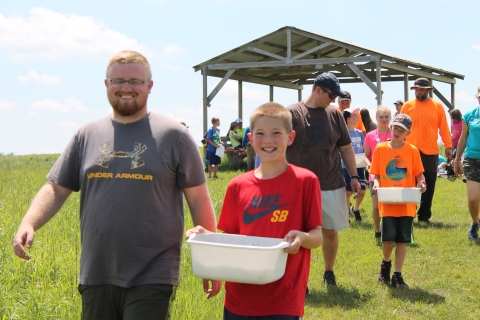 Adults and youth carry white tubs of lake water from a shelter building on a sunny summer day