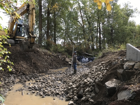 A man stands in a muddy, dewatered streambed looking up at an excavator perched on the bank.