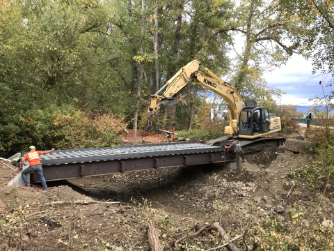 A flat, prefabricated bridge is lowered into place with an excavator.