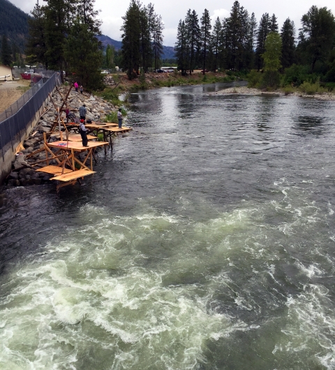 View over a rushing river with wooden platforms on the left bank and 3 men fishing from the platforms.