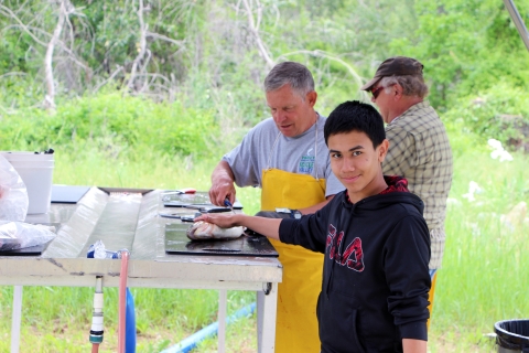 A young man stands with his hand on his fish, looking into the camera, while two men behind him work on cleaning the fish.