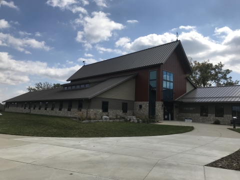 Looking towards the front entrance of a new facility with stone and red siding, large concrete walkway in foreground, small metal blue goose wind vanes on top of the facility against a blue sky with white puffy clouds