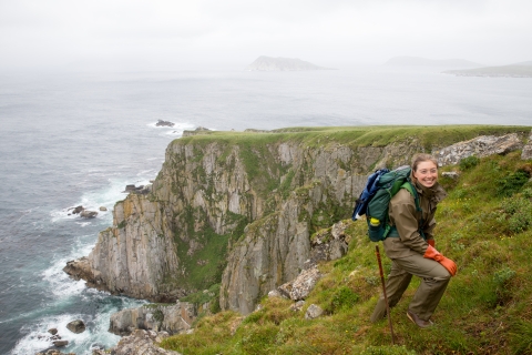 Woman wearing green rain suit, backpack, and orange gloves hikes up from a a survey site with cliff and ocean in the background.