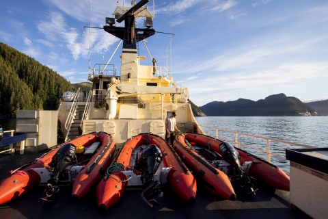 Three orange skiffs on the back deck of a large ship at sea.