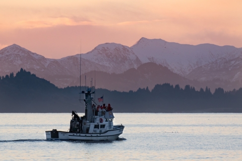 A white vessel with people standing on the flying bridge and purple and orange sunset over snowy mountains in background.