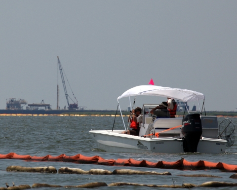 A biologist looks through binoculars from a boat near oil booms.