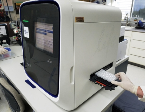 A large white qPCR machine has a visible screen on the front and a tray with multiple holes is being loaded into the side of the machine. 