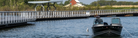 A fishing pier stretches into the river while an angler catches a fish from a boat nearby.