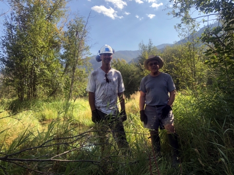 A tall man in hardhat, dirty t-shirt, and ear protection stands next to a shorter man in straw hat, shorts, and t-shirt, both wearing work gloves, in a field.