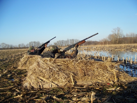 Two hunters in dirt camouflage bunker with shot guns raised to the sky. 