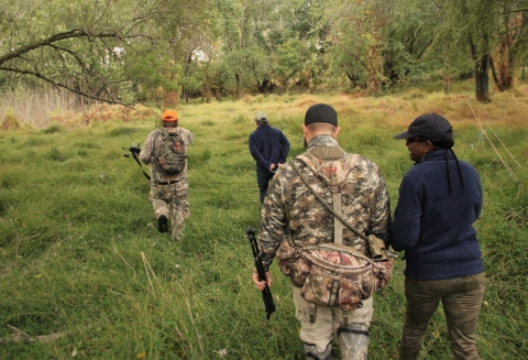 Four people -- two in hunting attire and two in regular attire -- walk across a grassy patch of forested area in a wildlife refuge