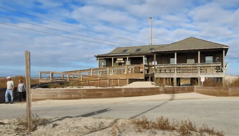 From left to right, a man and a womam walking away from a bulding. On the right: a front view of an earth tone building resting on piles raised five feet off the ground.