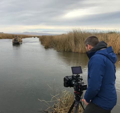 A man with his back to the camera using his camera on a tripod to shoot video of a small boat navigating a marshy waterway