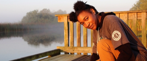 Young woman on dock in wetland wearing a hoodie emblazoned with the AmeriCorps logo